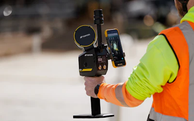 Construction worker in safety gear holding surveying equipment with smartphone display on job site