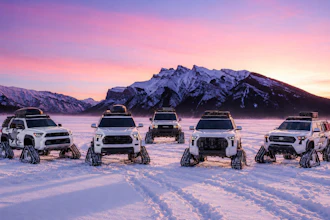 Five white Toyota pickup trucks and SUVs with tracks instead of wheels lined up on a frozen lake at sunset with snow-capped mountains in the background