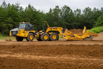 Yellow K-TEC motor grader with articulated scraper attachment working on dirt site surrounded by pine forest