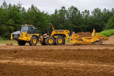 Yellow K-TEC motor grader with articulated scraper attachment working on dirt site surrounded by pine forest